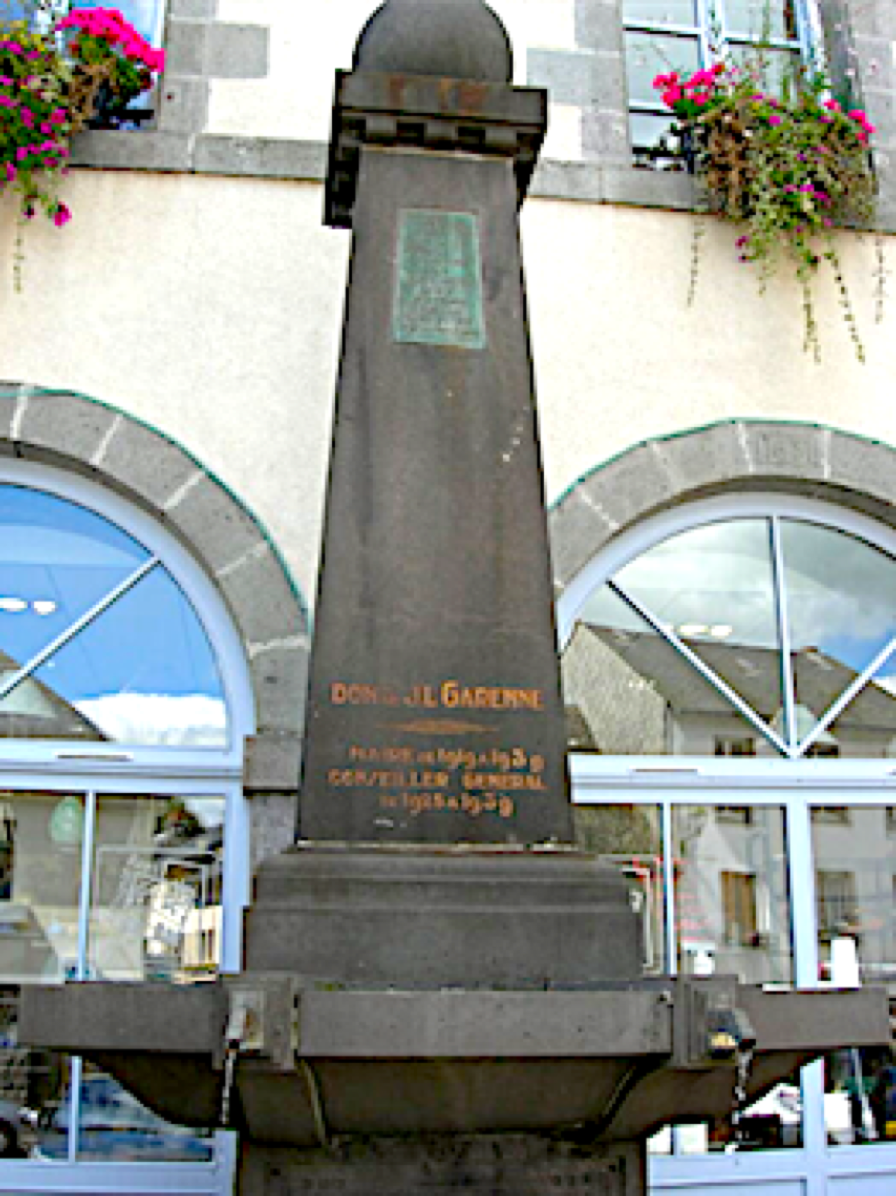 Monument en pierre sombre avec des inscriptions, devant un bâtiment avec des fenêtres en arc et des fleurs suspendues.