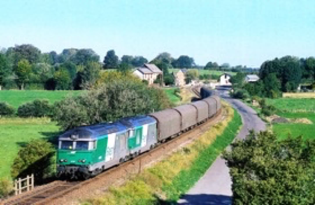 Train de marchandises vert et gris traversant un paysage rural avec des champs verts et quelques maisons à l'arrière-plan.