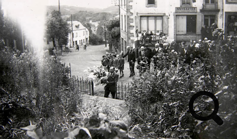 Une rue bordée de bâtiments avec un groupe de personnes rassemblées, des arbres et des buissons au premier plan.