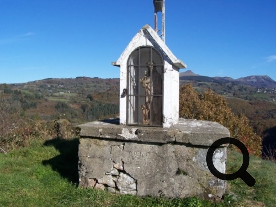 Petite structure en pierre avec une niche abritant une statue, située sur une colline avec un paysage de montagnes en arrière-plan.
