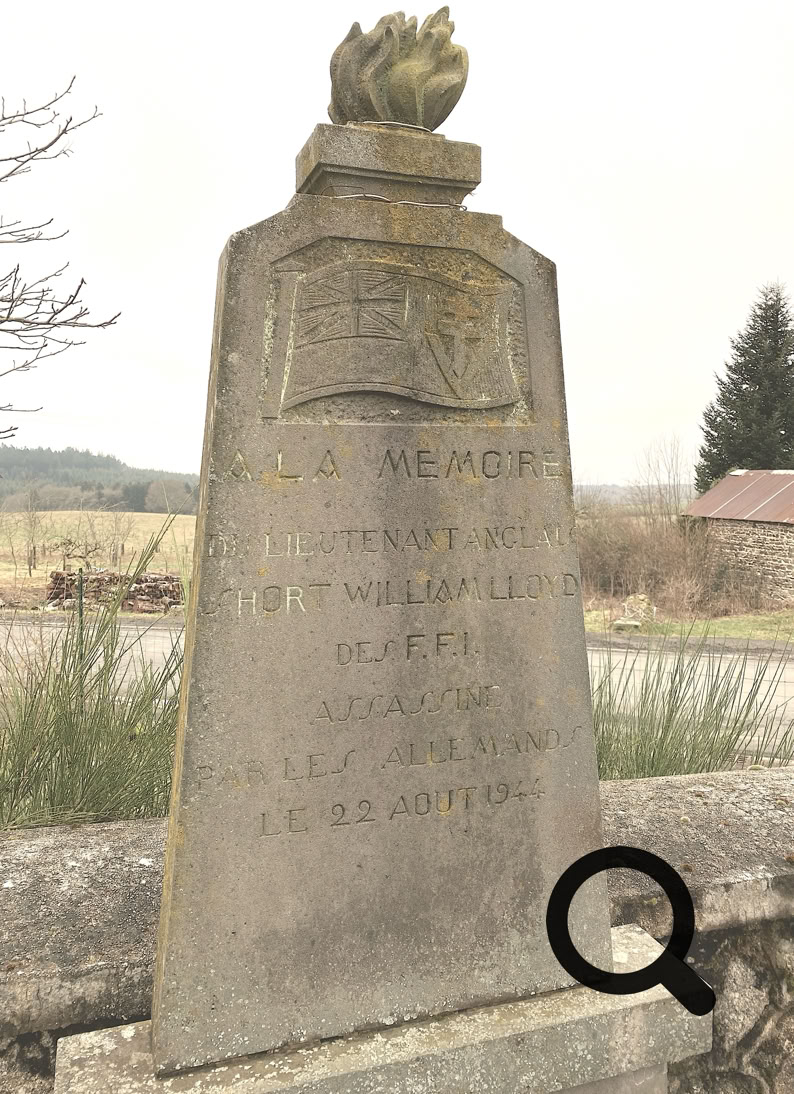 Monument en pierre avec des inscriptions, surmonté d'une sculpture en forme de flamme, situé à l'extérieur près d'une route et de la végétation.