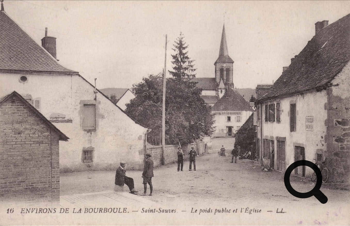 Rue de village avec des bâtiments anciens de chaque côté, quelques personnes marchant ou assises, et une église avec un clocher en arrière-plan.
