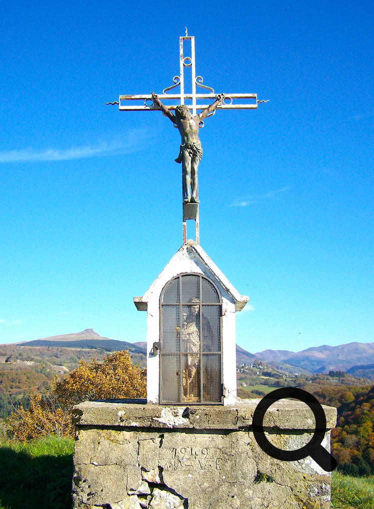 Croix en métal avec une statue sur un socle en pierre, paysage de montagnes en arrière-plan sous un ciel bleu.