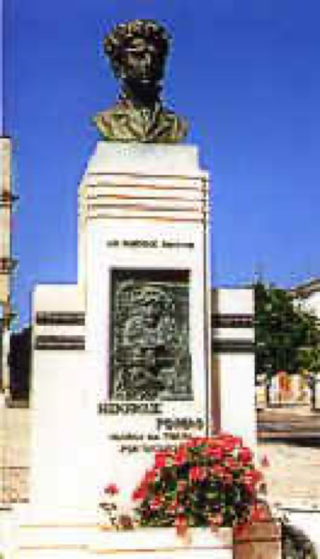 Buste en bronze de Henrique Pousao sur un socle en pierre avec une plaque sculptée, entouré de fleurs rouges, sous un ciel bleu.