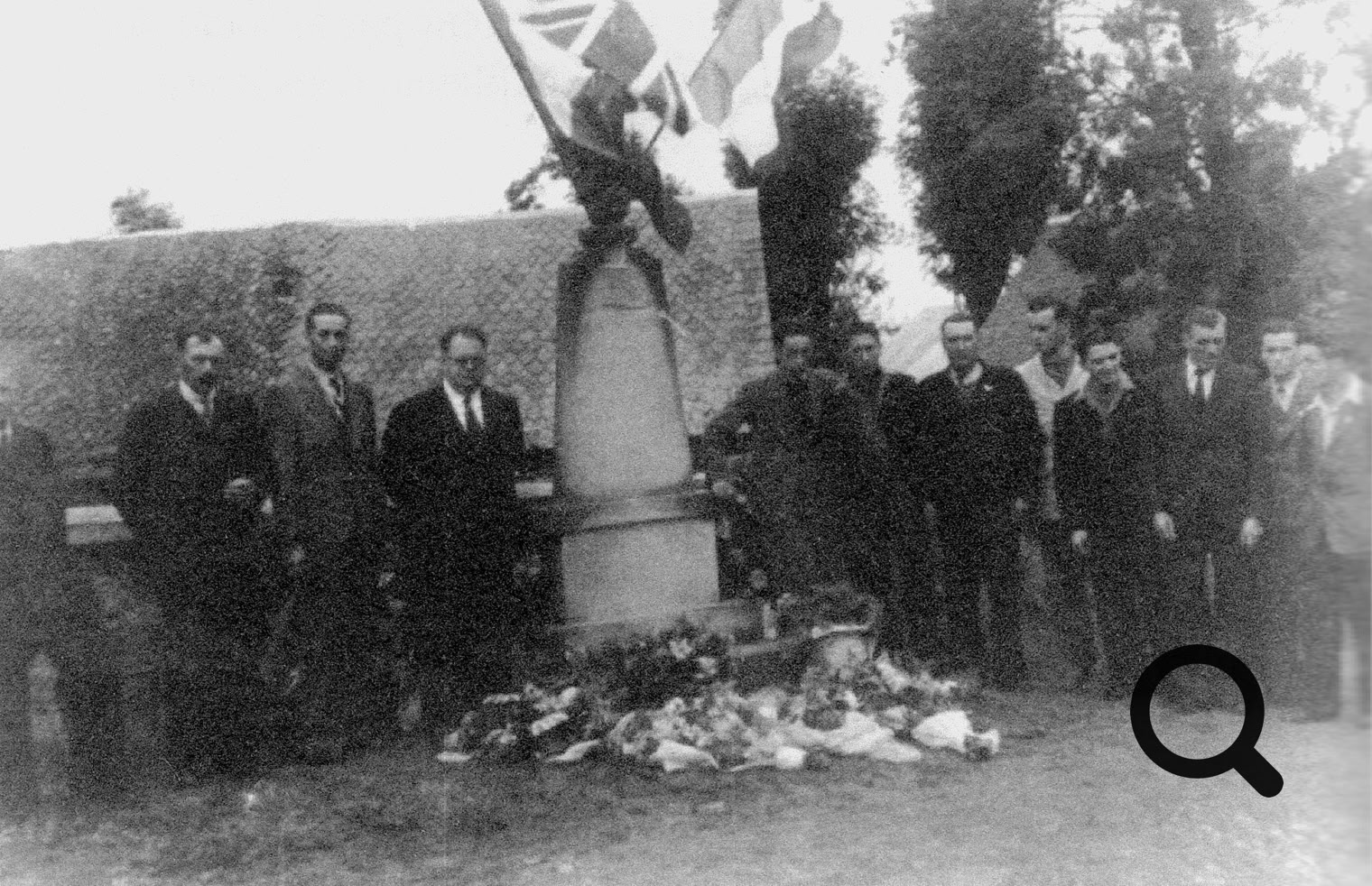 Un groupe de personnes posant devant un monument avec des drapeaux et des fleurs déposées à la base.