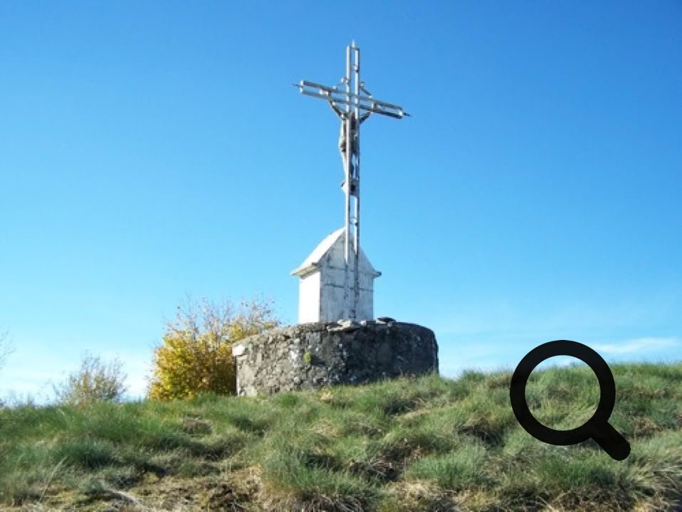 Croix en métal sur un socle en pierre, située sur une colline herbeuse sous un ciel bleu.
